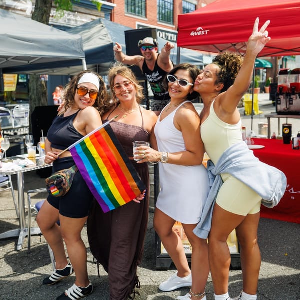 Four smiling women pose with a rainbow flag at a lively outdoor event with a DJ.