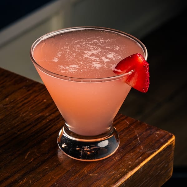 Pink cocktail in a martini glass, garnished with a strawberry slice, on a wooden surface.