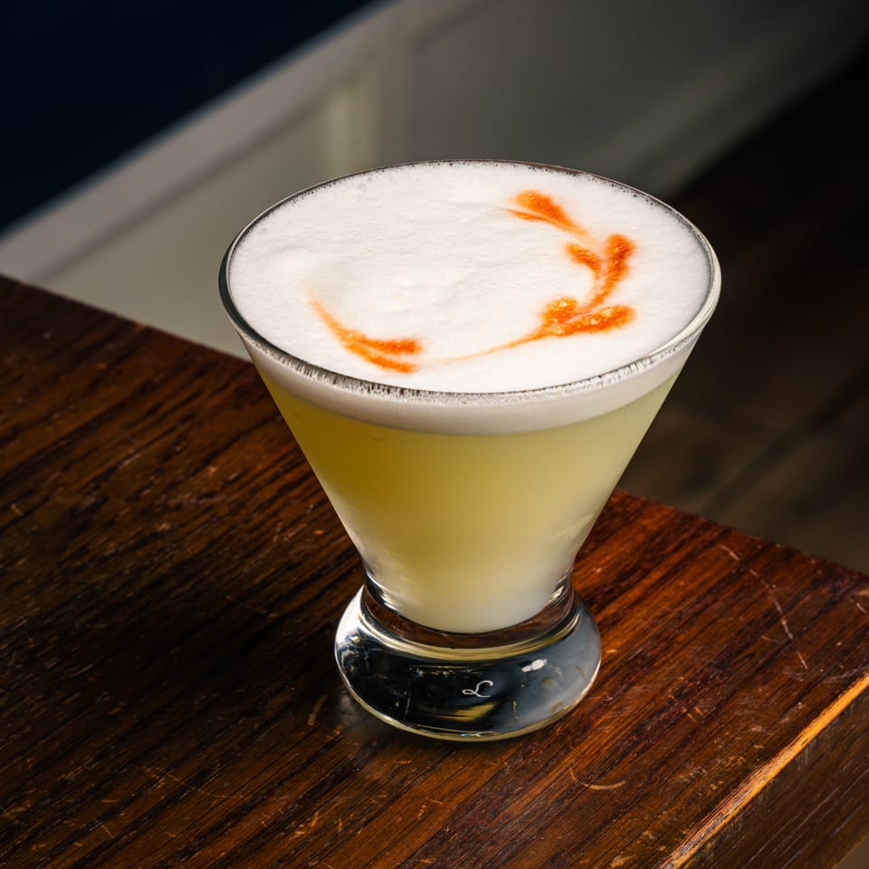 Peruvian Pisco Sour cocktail with a foamy top and an orange garnish, on a wooden surface.