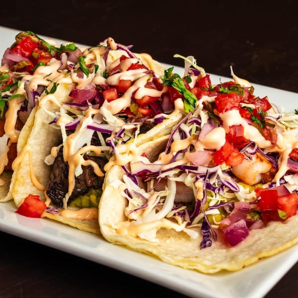 Three Peruvian tacos on a white plate, filled with meat, shrimp, cabbage, salsa, and a creamy sauce.