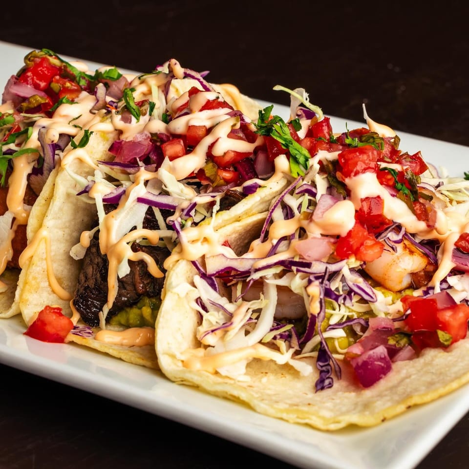 Three Peruvian tacos on a white plate, filled with meat, shrimp, cabbage, salsa, and a creamy sauce.