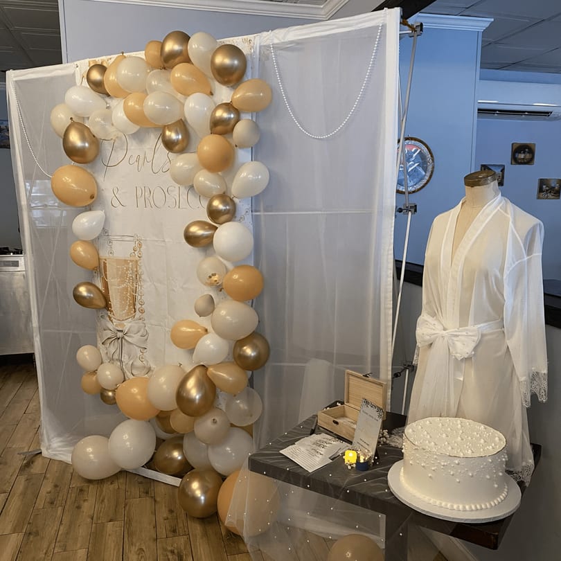 Elegant 'Pearls & Prosecco' balloon backdrop, white cake, and bridal robe display for an event.