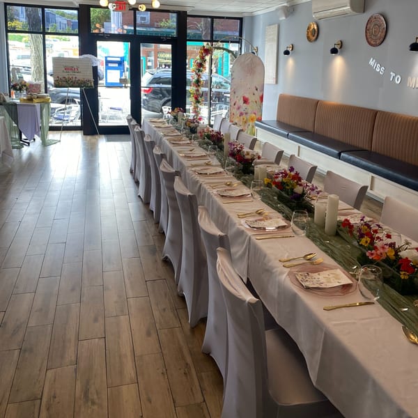 Long dining table set for a bridal shower with floral decor, white chairs, and street view.