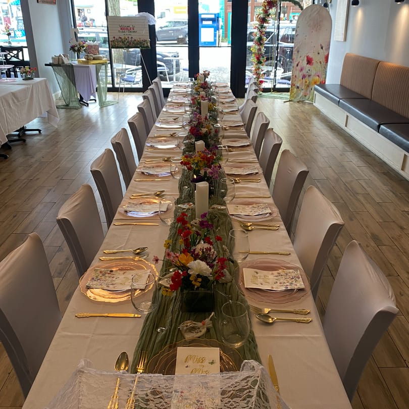Long dining table set for a bridal shower with floral centerpieces, gold cutlery, and white chair covers.