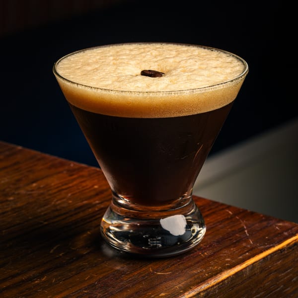 A dark espresso martini with a thick foam and a single coffee bean on a wooden table.