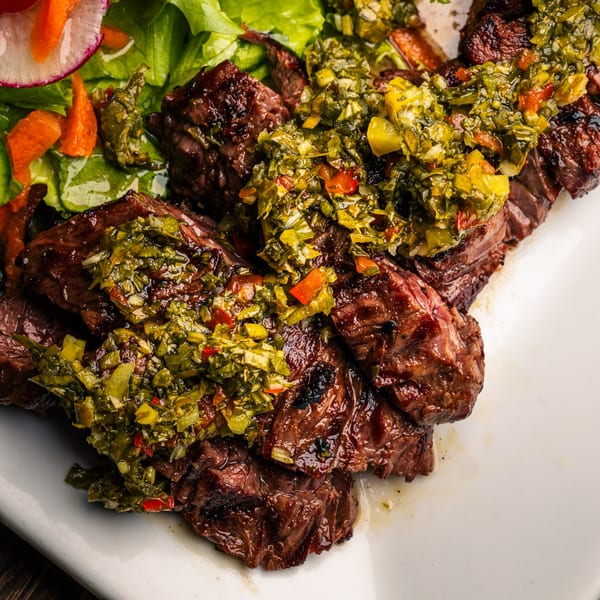 Grilled steak with vibrant green chimichurri sauce and a side salad on a white plate.