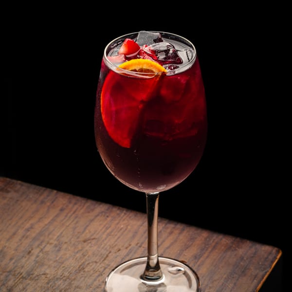 A glass of Maura's Kitchen's red sangria with ice, orange slice, and strawberries on a wooden table.