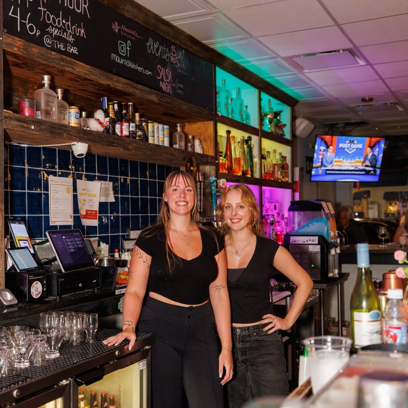 Two smiling women stand behind a colorful bar with bottles, glasses, and a chalkboard menu.