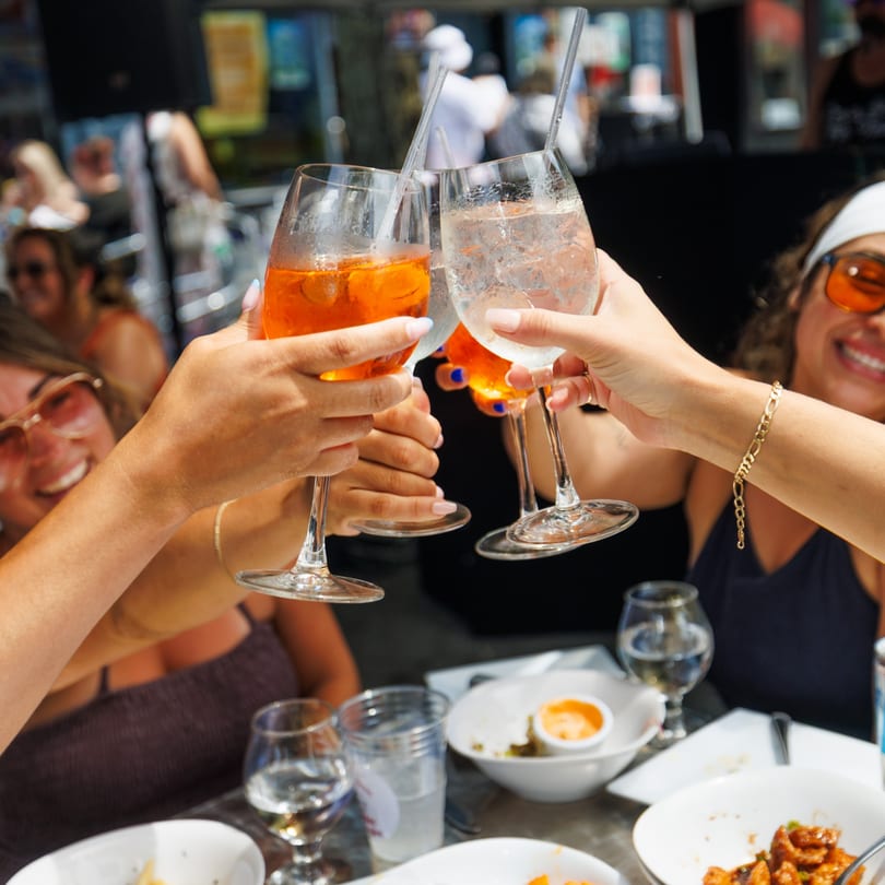 Close-up of friends toasting with orange cocktails and water at a vibrant outdoor restaurant.